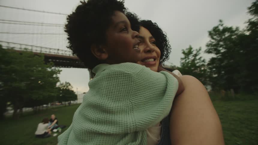 The video shows a woman holding a young child in a parklike setting with the iconic brooklyn bridge visible in the background they are both looking towards the bridge suggesting they are enjoying the view together