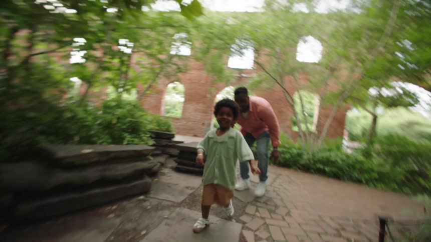The child runs joyfully along a brick path through a garden with the father playfully chasing behind