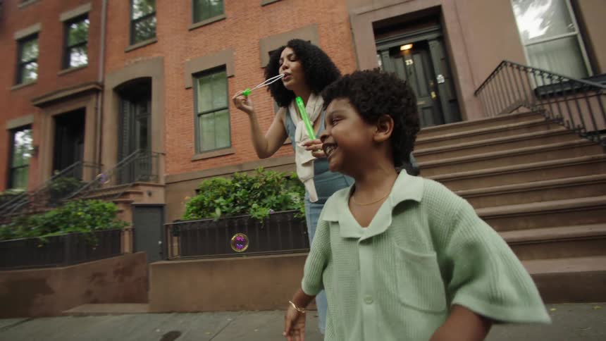 A family plays with bubbles on a quiet sidewalk in front of classic redbrick brownstones with the child joyfully reaching for bubbles as both parents engage