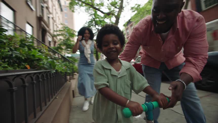 A father guiding his son on a scooter along the sidewalk with the mother walking behind and watching the fun moment at midday