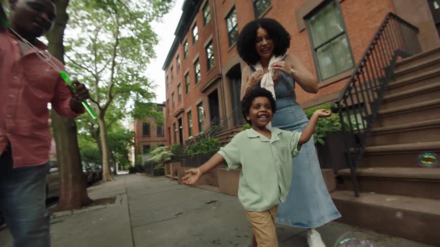 A joyful family blows bubbles and plays together on a treelined sidewalk in front of redbrick brownstones on a sunny day