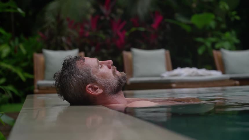A man with graying hair and a beard relaxes in a tranquil pool leaning his head back against the edge while surrounded by lush greenery and lounge chairs
