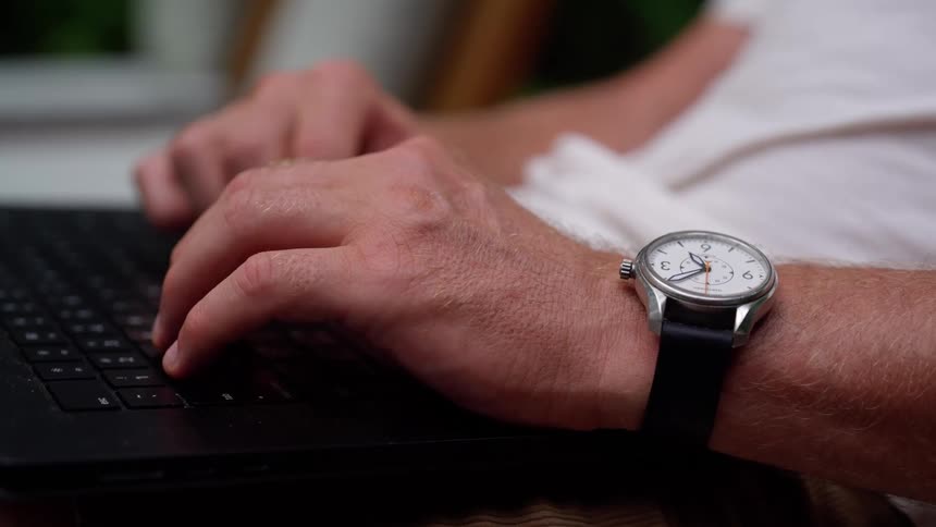 A man wearing a watch seated and typing on a laptop