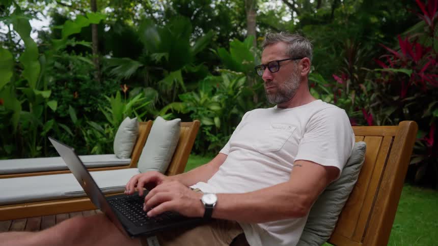A middleaged man sitting on an outdoor pool chair typing on his laptop placed on his lap at midday