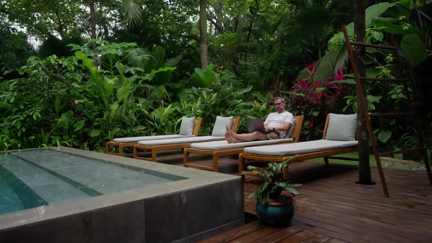 Older man sits on a lounge chair near an outdoor pool and works on a laptop