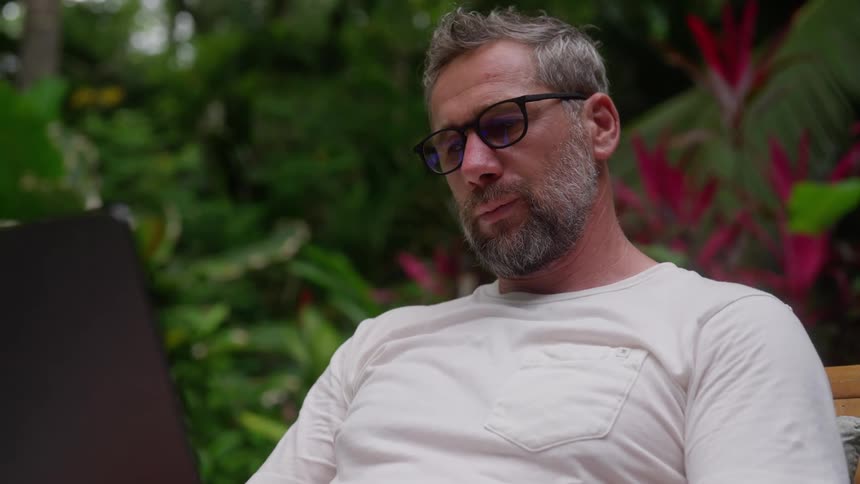 Seated man wearing glasses interacting with laptop in an outdoor setting with plants in the background