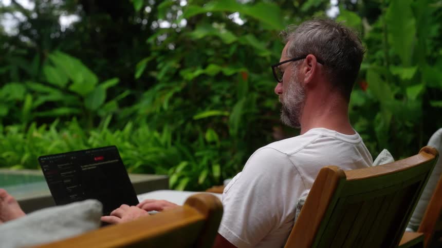 Older man sits in a lounge chair at poolside and types on a laptop with a brief pause to check his wristwatch