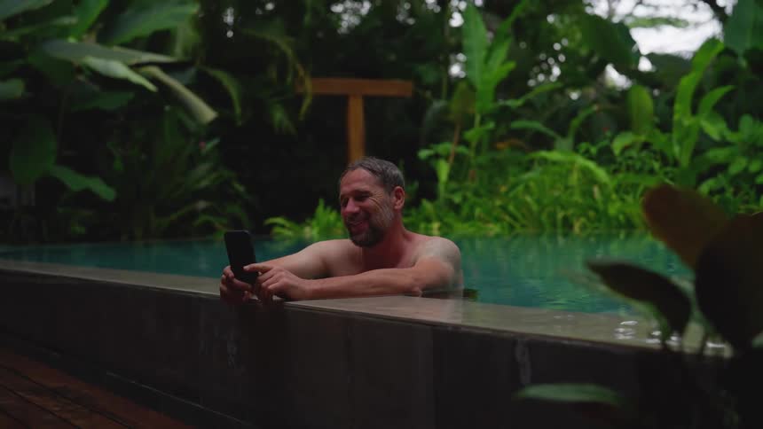 A man in a pool using his mobile device with a smile on his face at midday