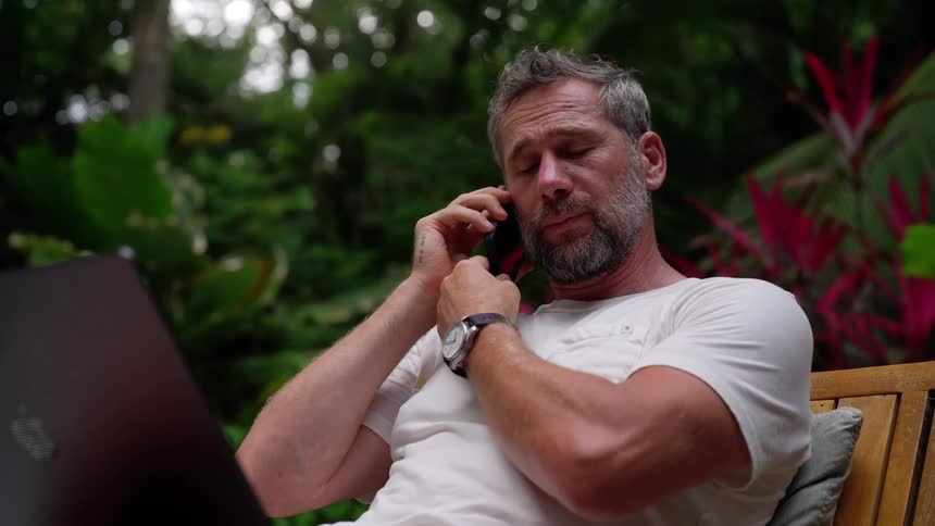 A man takes a phone call while seating outdoors with his laptop on a lounge chair during daytime