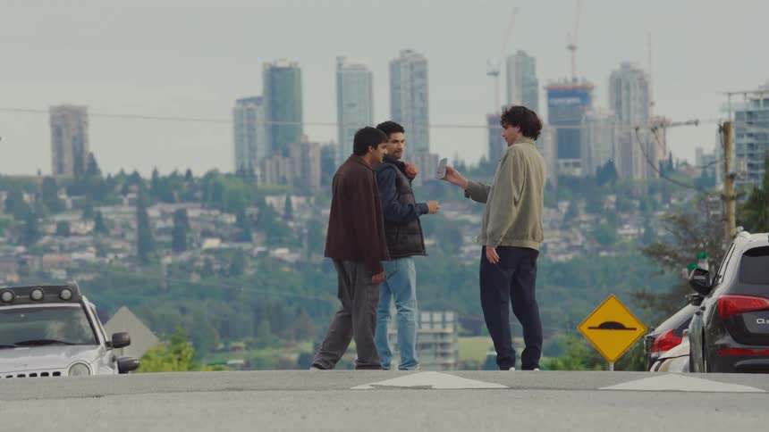 Three men stand and talk in the middle of a street with cars parked nearby overlooking a city skyline with tall buildings and construction cranes in the distance