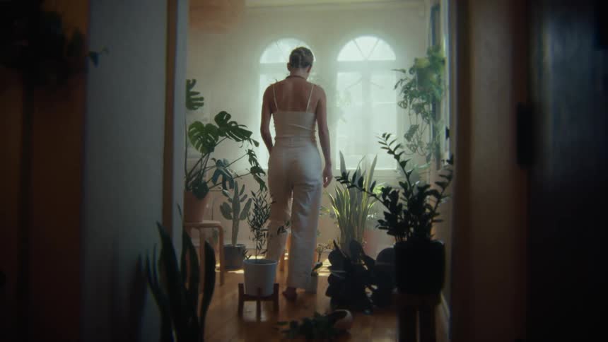 A young woman walks into an indoor garden with potted plants during daytime and sits on a wooden stool