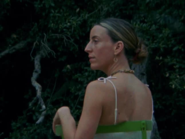 A woman with her hair tied back sits on a green chair looking off to the side with dark foliage and pink flowers in the background