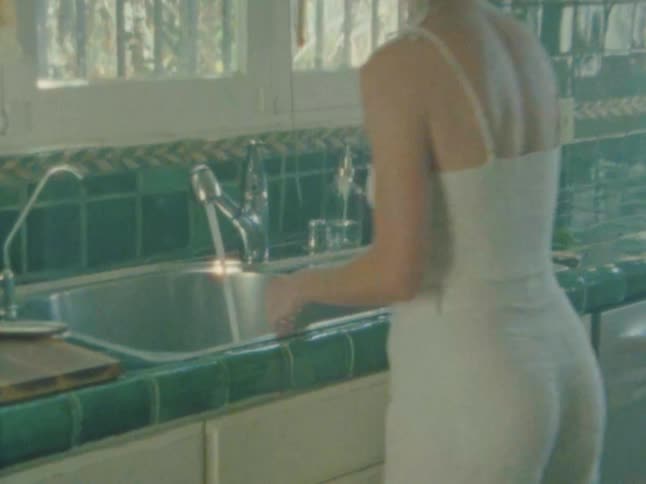 A woman in a white tank top is seen from behind leaning over a kitchen sink washing her hands with her back to the camera with green tiled countertops and a window letting in bright sunlight