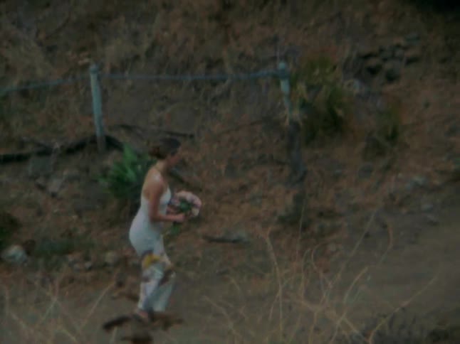 Woman carrying a bouquet walks along a dirt path paralleling a fence
