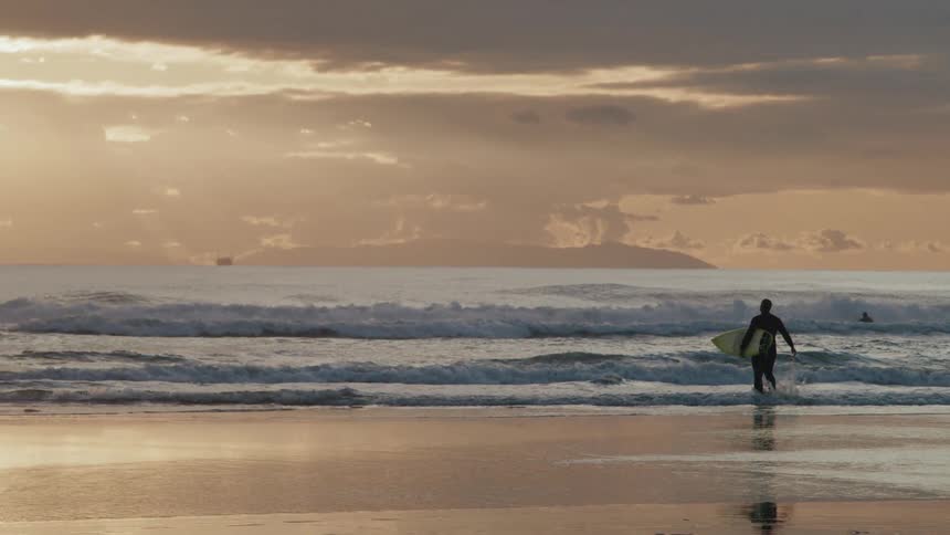 A lone surfer silhouetted against the setting sun stands in the shallow water at the edge of the ocean holding a surfboard and looking out at the rolling waves under a cloudy orange hued sky