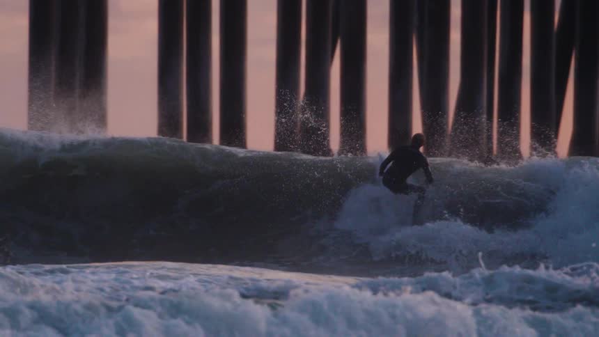 A surfer in a dark wetsuit rides a wave with a pier in the background the surfer performs an aerial maneuver launching off the wave with the surfboard