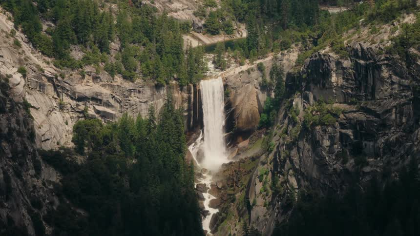 The video presents a grand view of a powerful waterfall cascading down a sheer rock face surrounded by rugged treecovered mountains the water plunges into a rocky basin below with a river flowing away into the distance