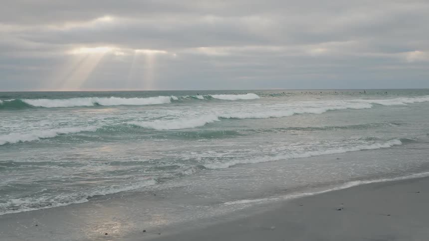The video shows an expansive view of the ocean under a cloudy sky with rays of sunlight breaking through the clouds and illuminating parts of the water waves are visible rolling towards the shore and several surfers can be seen in the distance waiting