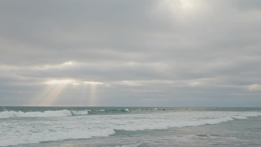 The video shows an expansive view of the ocean under a cloudy sky with rays of sunlight breaking through the clouds and illuminating parts of the water waves are visible rolling towards the shore and several surfers can be seen in the distance waiting