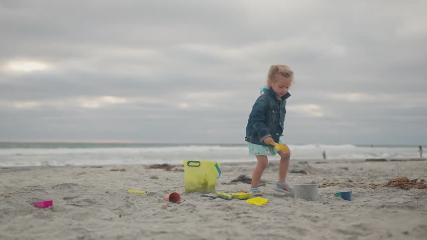 A child in a blue jacket and light dress plays on a sandy beach pouring sand into a yellow bucket surrounded by scattered beach toys