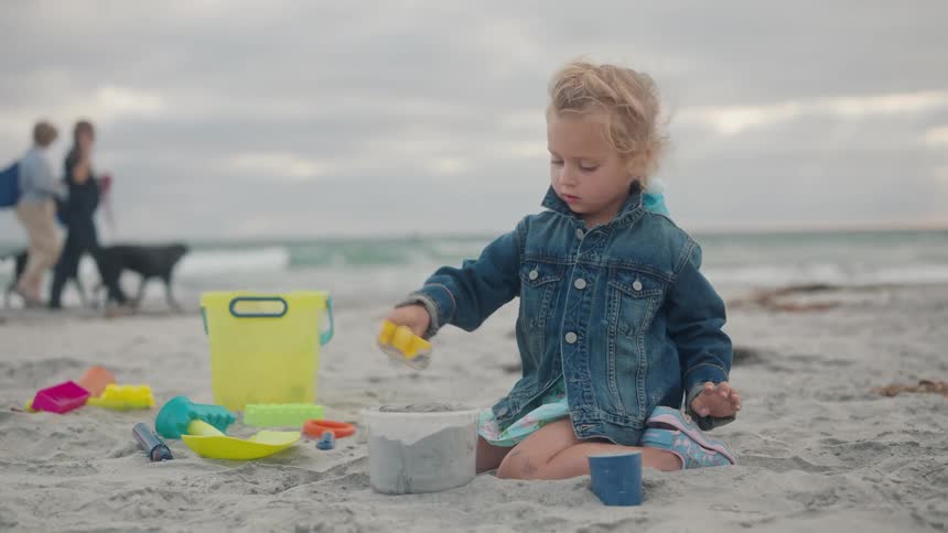 Blonde girl with jeans jacket playing with sand in the beach and two women walking with dogs at the background