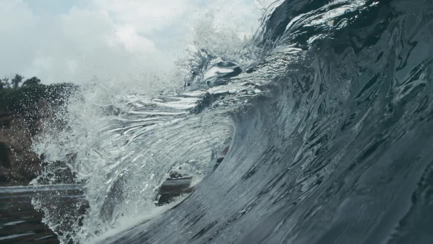 A large ocean wave curls near a rocky coastline sending spray upward as it passes in front of vegetationcovered cliffs