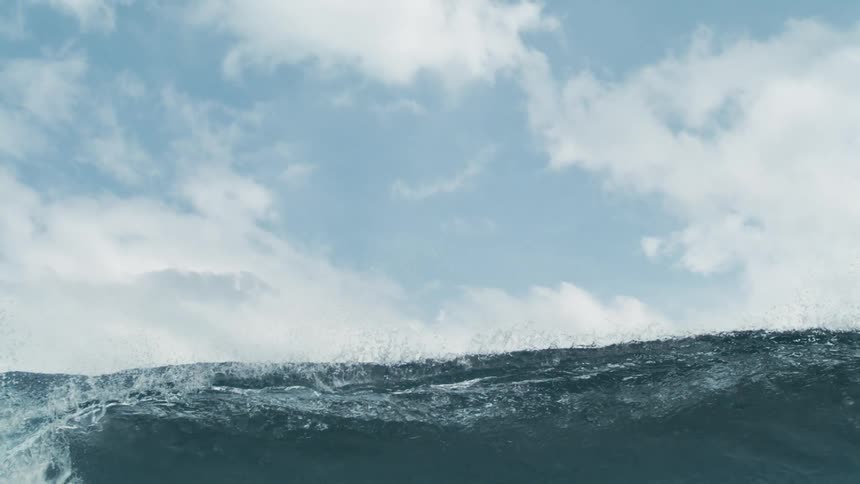 A large wave crashes in the ocean under white fluffy clouds