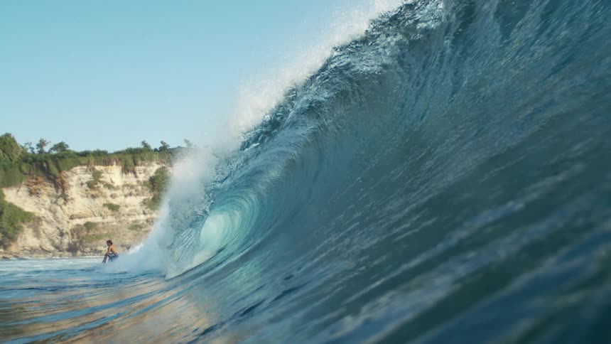 A surfer glides along a crystalblue wave near rugged cliffs under a clear sky