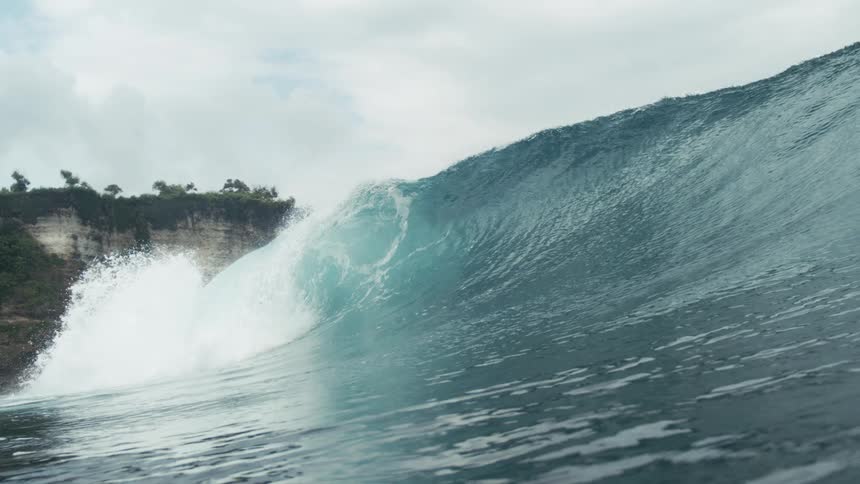 A large wave crashes near a coastal cliff on a cloudy day