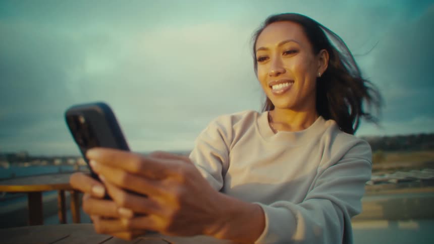 A smiling asian woman is holding a smartphone looking up and away from the camera with a cloudy sky and distant water visible behind her