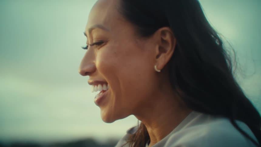 A smiling asian woman is looking down at her phone holding it with both hands with a bright hazy sky and water in the background
