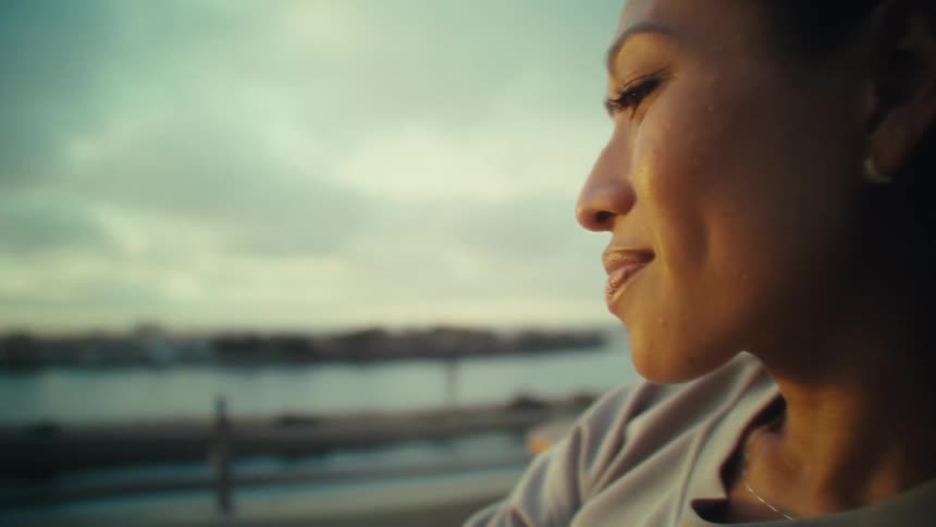 An asian woman is looking at her phone with a calm expression silhouetted against a serene waterfront sunset