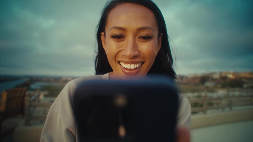A woman beams joyfully while looking at her phone standing on a rooftop with a scenic coastal backdrop
