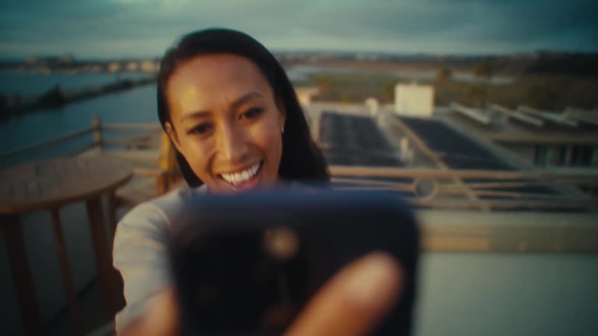 A young woman takes selfies with a mobile phone while standing on a building during daytime