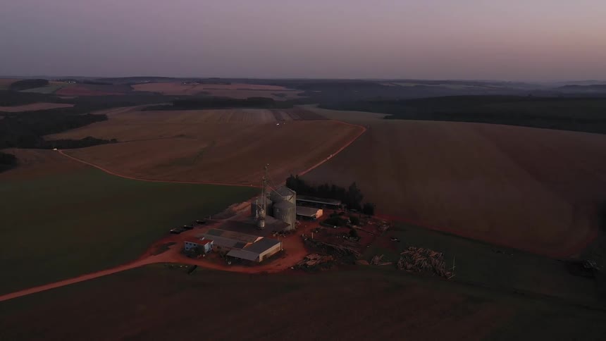 The video shows an aerial view of a vast agricultural landscape at what appears to be dawn or dusk with fields of varying shades of brown and green in the foreground a farm complex is visible featuring several tall silos and other smaller buildings