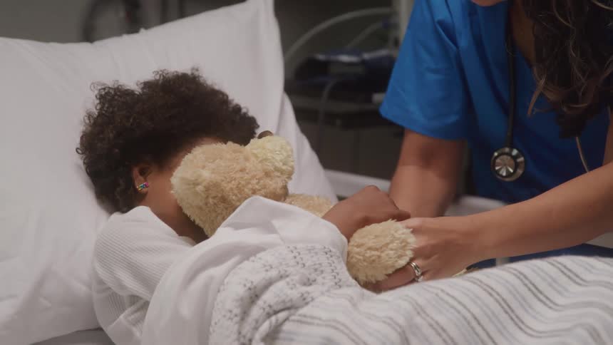 A nurturing nurse offers comfort to a young child who is resting in bed and embracing a teddy bear