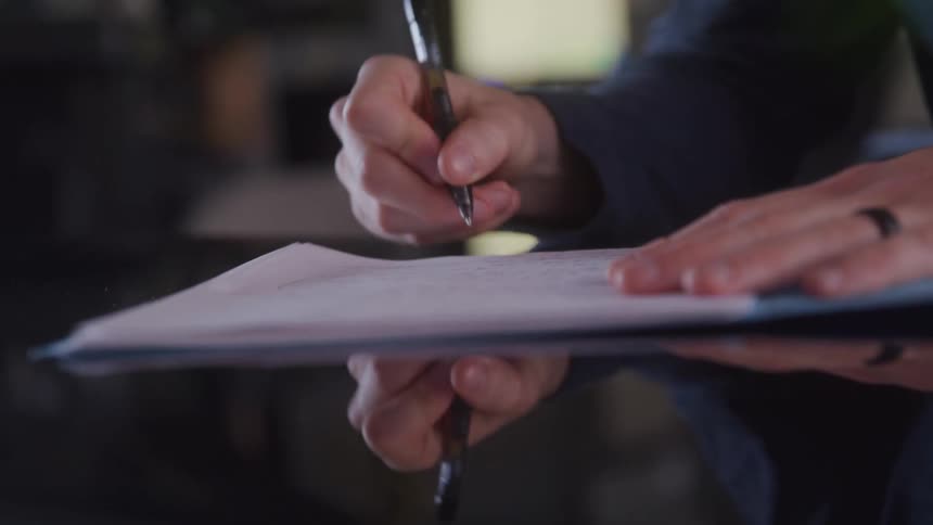 A person signs a document on a clipboard with a firm grip and focused posture