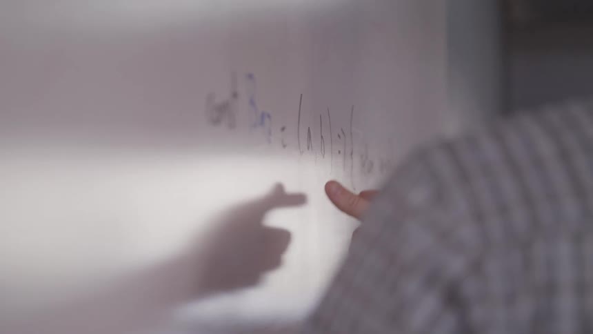A close up of a hand pointing to a whiteboard filled with writing as the person gestures mid explanation