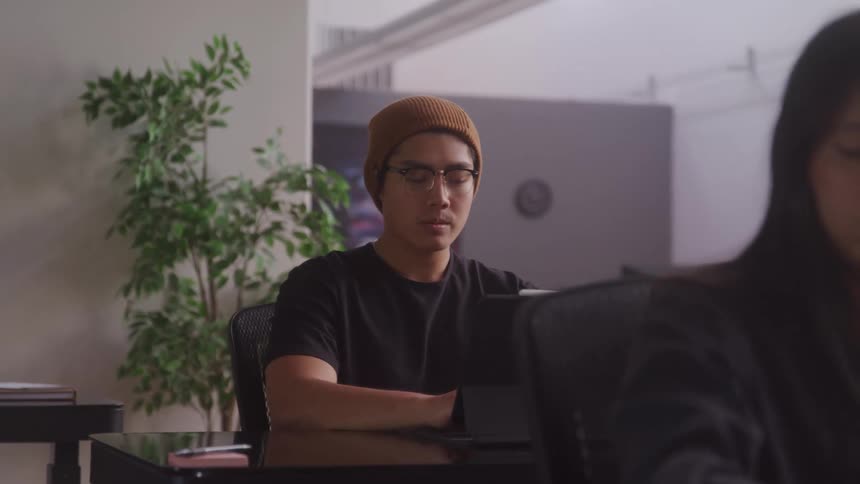 A man wearing a beanie and glasses is sitting at a desk with a serious and attentive expression and a girl in front making notes