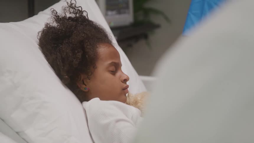 Young girl lies in hospital bed holding toy as healthcare worker cares beside