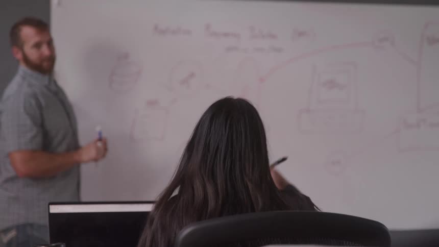 A student raises her hand during a lesson while a teacher looks on at a whiteboard