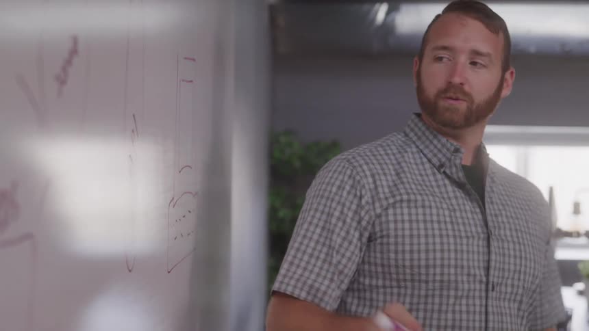 A man with a beard and a plaid shirt is actively drawing or writing on a white board with a red marker he periodically pauses to look away from the board seemingly contemplating his next mark or engaging with someone offcamera