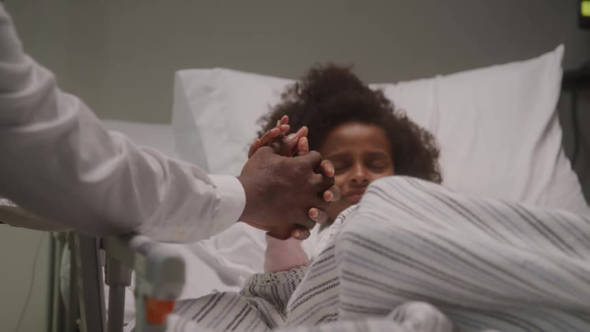 A person is seen in a hospital room holding the hand of another person who is lying in a bed the person in bed appears to be a child with curly hair looking up at the other individual