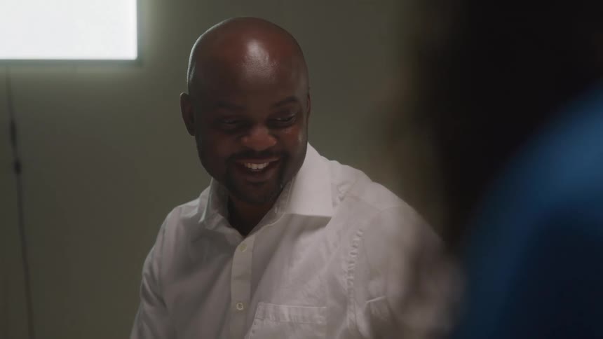 A bald darkskinned man in a white collared shirt smiles and looks to his right with a blurred blue figure in the foreground and a window in the background
