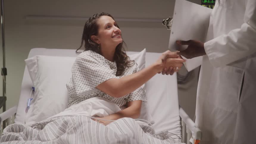 A female patient is lying in a hospital bed smiling as she shakes hands with a doctor standing beside her medical equipment with illuminated screens displaying vital signs is visible in the background
