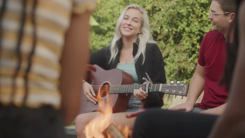 Woman plays guitar while sitting outdoors in front of a campfire surrounded by other people