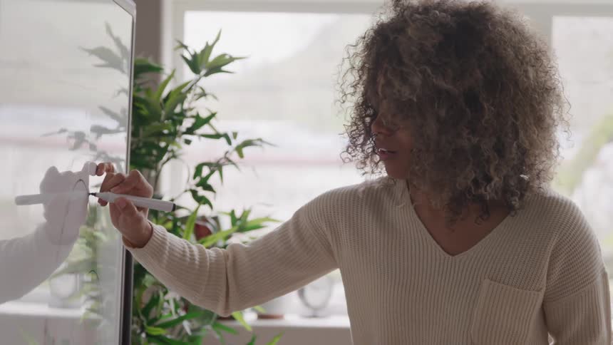 A woman with curly hair is enthusiastically writing on a whiteboard with a marker actively gesturing with her other hand as if explaining something she is positioned indoors next to a window with a plant visible behind her