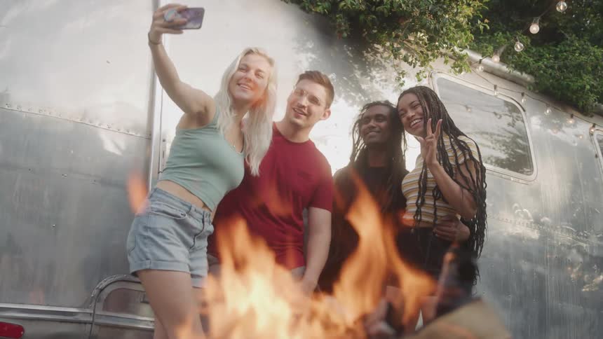 Four friends beside an rv taking a selfie with a burning fire in front of them at midday