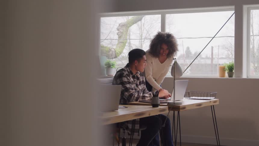 The video shows a bright office space with large windows revealing trees outside where two colleagues are working together one person is seated at a desk using a laptop while the other leans over and points at the screen engaging in discussion