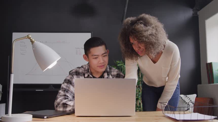 Two colleagues are collaborating at a desk with one person seated in front of a laptop and the other leaning over to point at the screen they both appear engaged in discussion looking at the laptop screen together